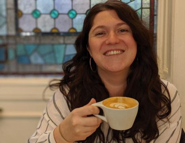 Emmie Harrison-West holding a coffee while sitting indoors - there's a wall with a stained glass window behind her and she's smiling at the camera.