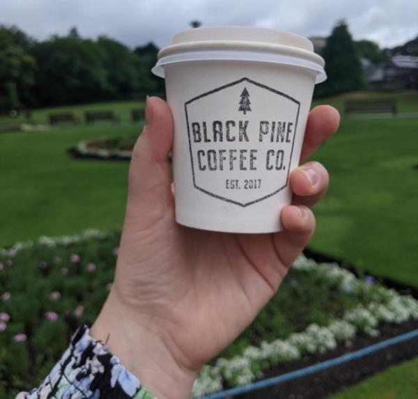 Emmie Harrison-West's hand holding a takeaway coffee cup that reads 'BLACK PINE COFFEE CO.' – a park with green grass/flowerbeds in the background.