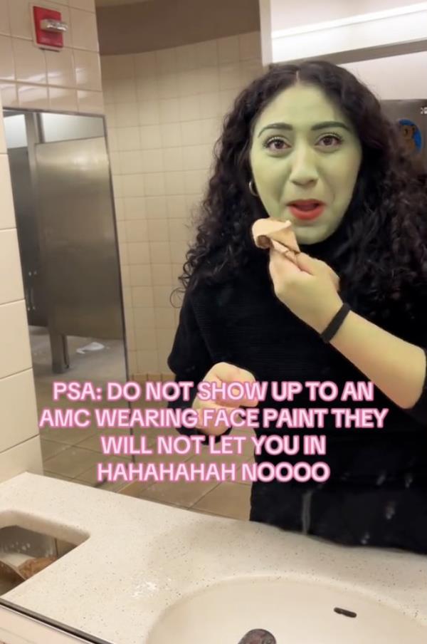 A woman with curly hair and her face painted green in support of Wicked, being asked to remove the face paint before a movie screening at AMC Theater.