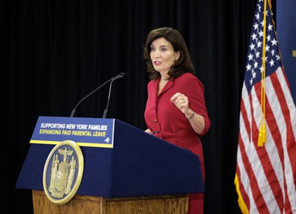 Kathy Hochul speaking at a podium in front of a black background and American flag with 