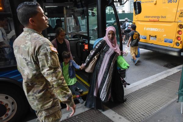 A woman with a hijab getting off a bus while holding a child's hand while a member of the Natio<em></em>nal Guard stands in camo on an NYC street.