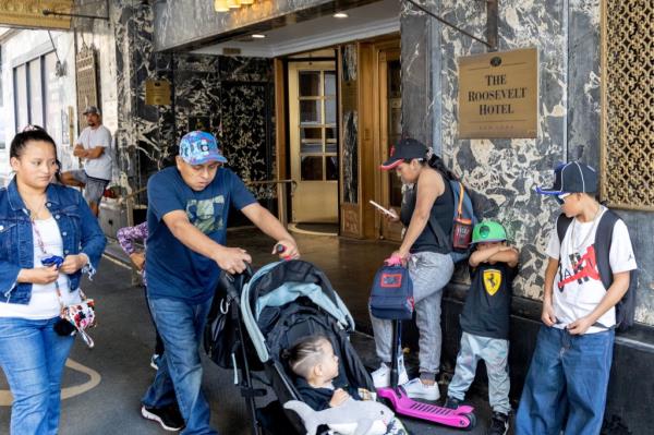 A migrant family with a stroller in front of the Roosevelt Hotel in NYC with several people leaning against a wall.