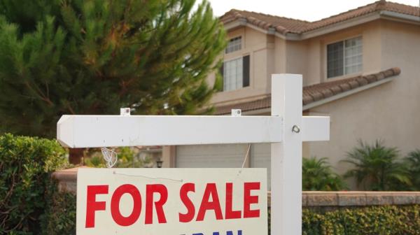 A for sale sign is posted outside a tan home with a tree.