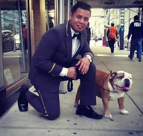 Stephen Bruno in doorman uniforman, kneeling on the sidewalk next to a bulldog. 